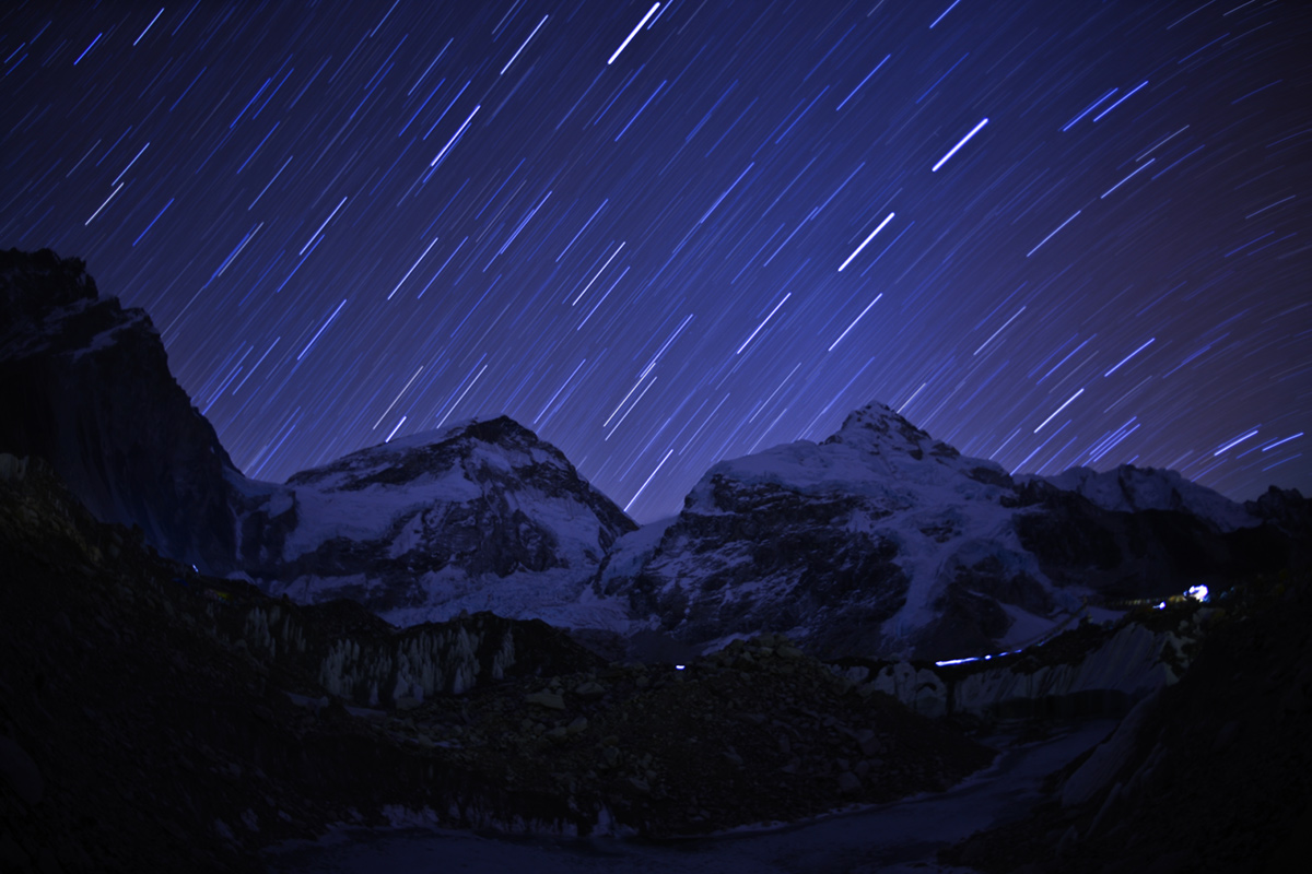 Stars wheel above the mountains and an illuminated camp