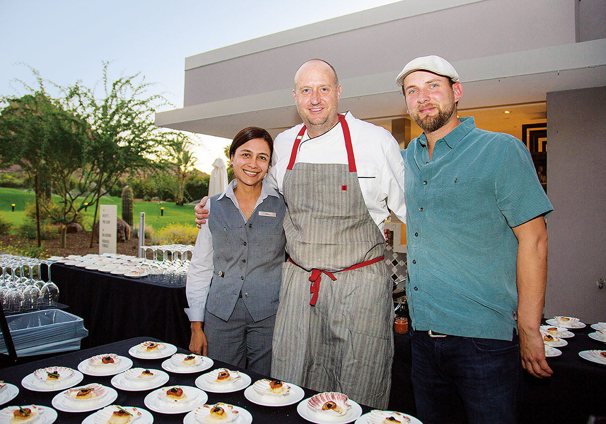 Chef Walter Sterling poses with a man and woman behind a catering table covered with food at outdoor event