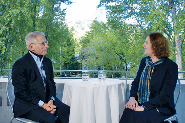 During the interview, Emory President Claire E. Sterk sits across a small table from Anthony Fauci, on a balcony with the leafy trees of Aspen, Colorado, in the background. 