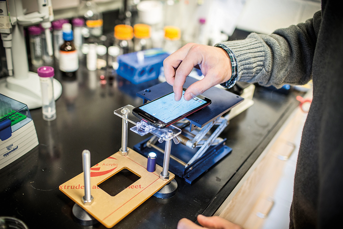 A hand manipulating a smart phone in a lab environment. The phone is mounted on a stand made of clear lucite and metal.