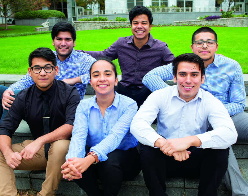 Six students pose smiling-five men plus a woman in the center-sitting on steps with a green lawn behind them.