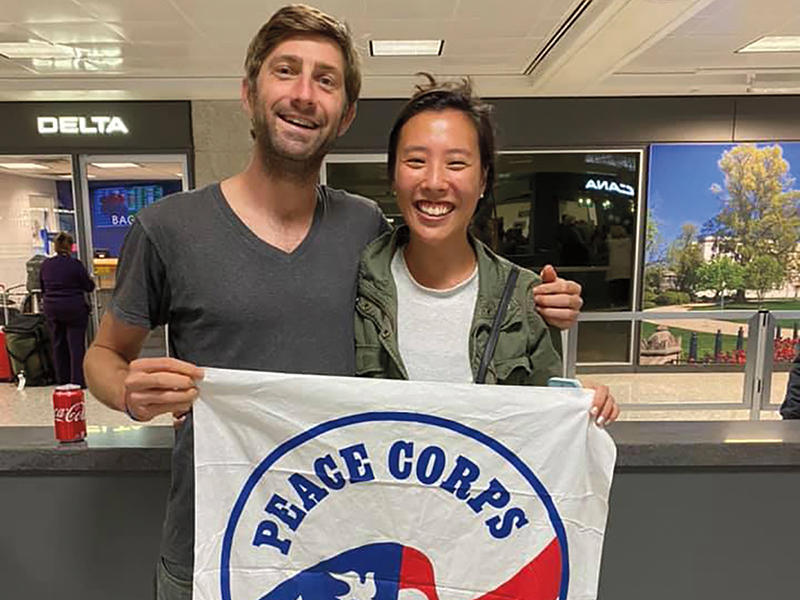 A man and a woman holding a Peace Corps flag stand together in an airport.